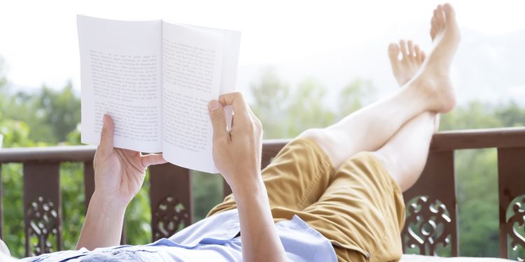 Photo of young man reading a book lying on soft mattress in relaxing bed at terrace